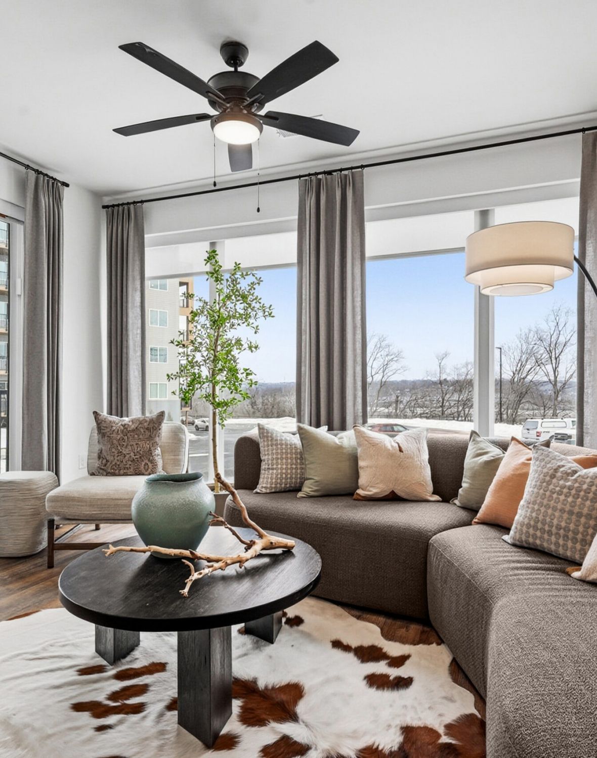 Modern living room with large windows, brown curved sofa, round table, and neutral decor accents.