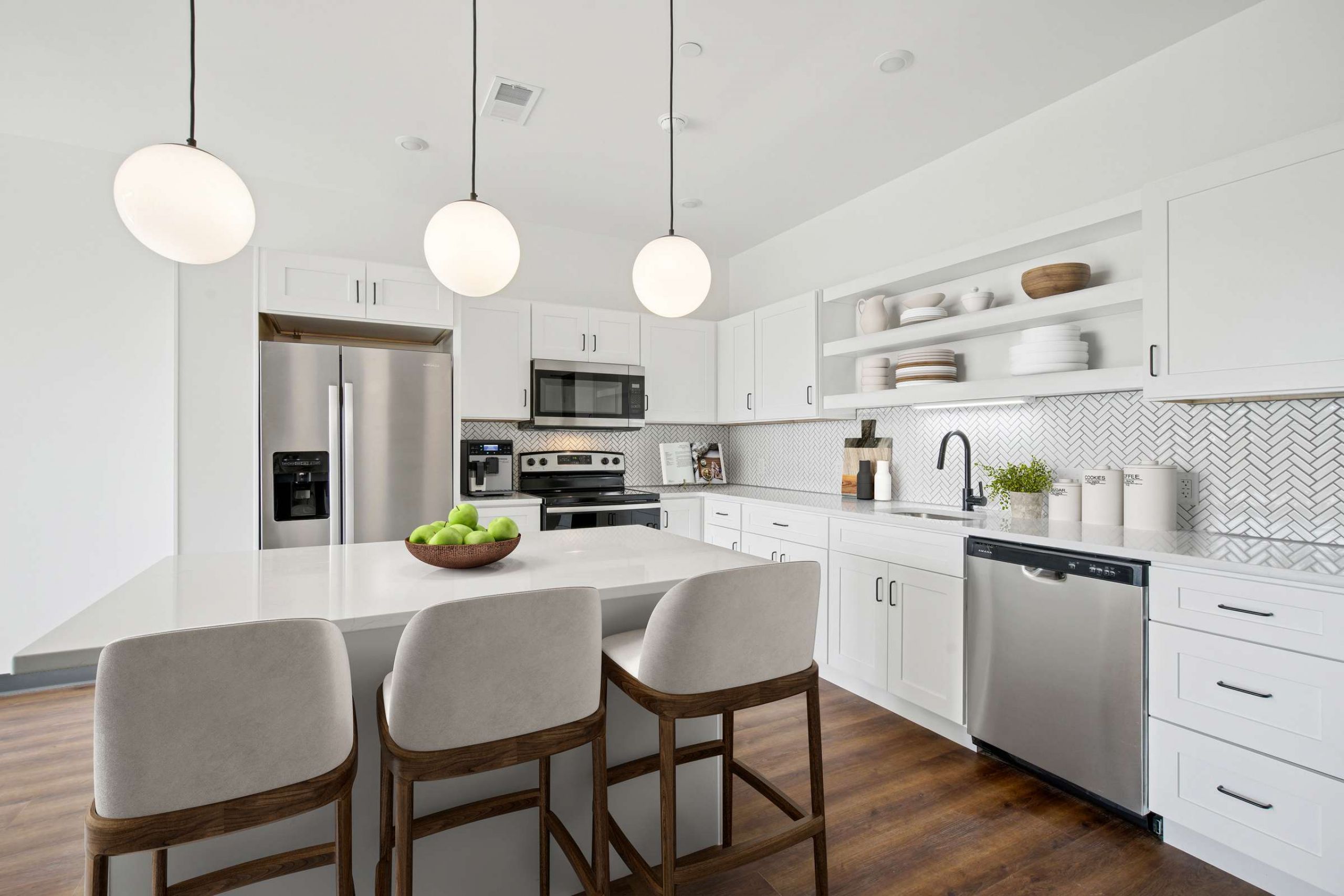 Modern white kitchen with island, three chairs, pendant lights, stainless steel appliances, and open shelves.