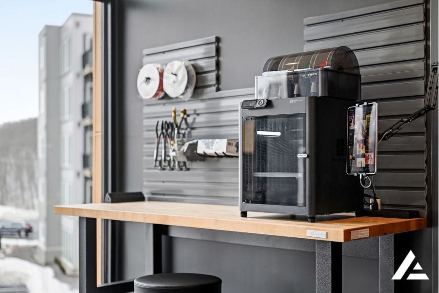 A 3D printer sits on a wooden workbench by a window, with tools and supplies hanging on the wall behind it.