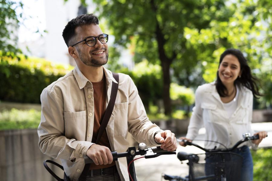 A smiling man and woman walk bikes outdoors on a sunny day, surrounded by greenery.