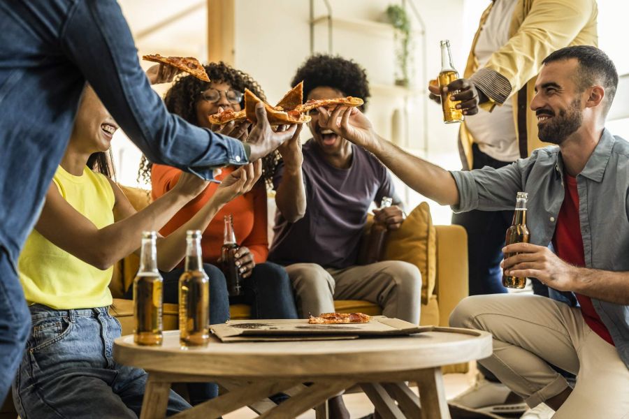 Friends laughing, eating pizza, and holding beer bottles together in a cozy living room setting.