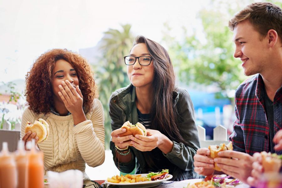 Three friends sit outdoors, laughing and eating burgers and fries at a table.