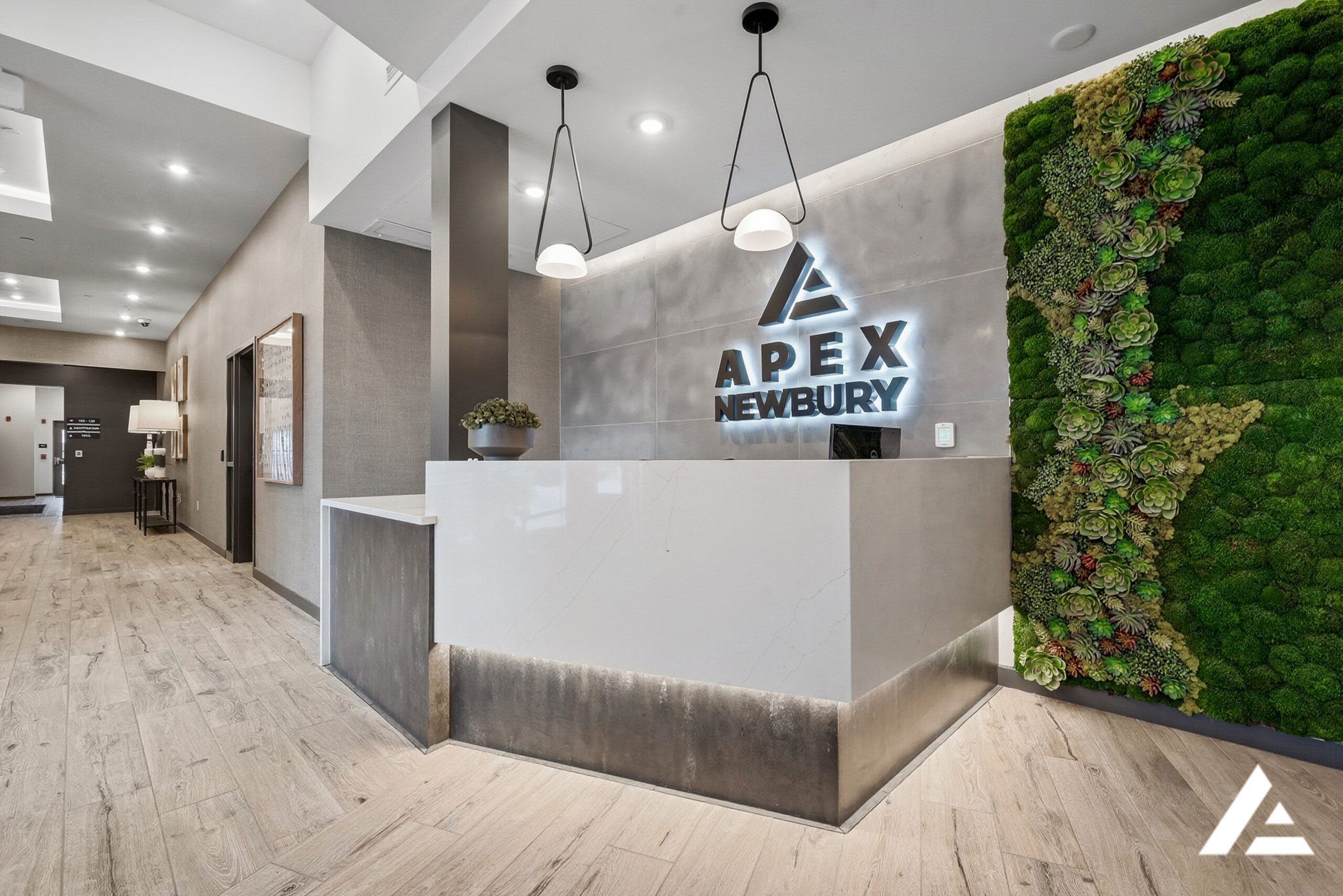 Modern reception desk with "Apex Newbury" sign, pendant lights, and a vertical green plant wall.