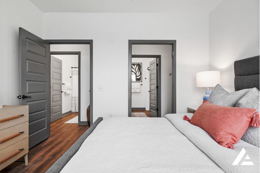 Modern bedroom with a neatly made bed, a dresser, and two open doors showing bathrooms and closets.