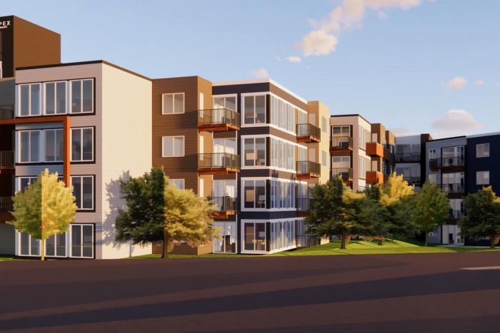 Modern apartment complex with large windows and balconies, surrounded by trees and a clear sky.